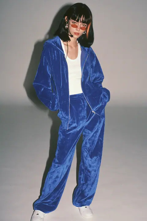 full-length shot, head to toe with the feet in frame, of a young Caucasian American woman with pale skin and a glossy black blunt bob with heavy straight bangs and one clay-orange streak, a thin black headset…