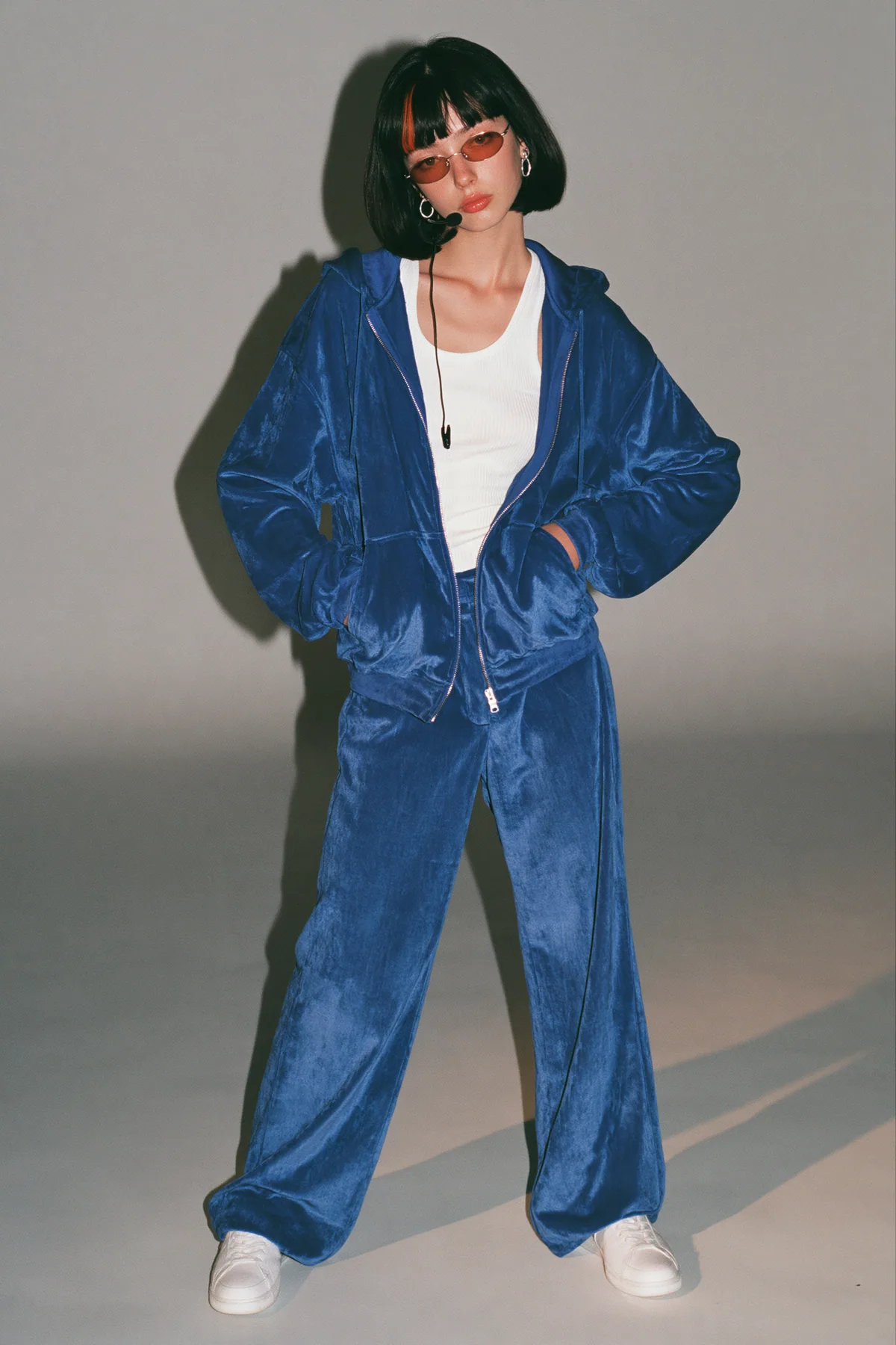 full-length shot, head to toe with the feet in frame, of a young Caucasian American woman with pale skin and a glossy black blunt bob with heavy straight bangs and one clay-orange streak, a thin black headset…