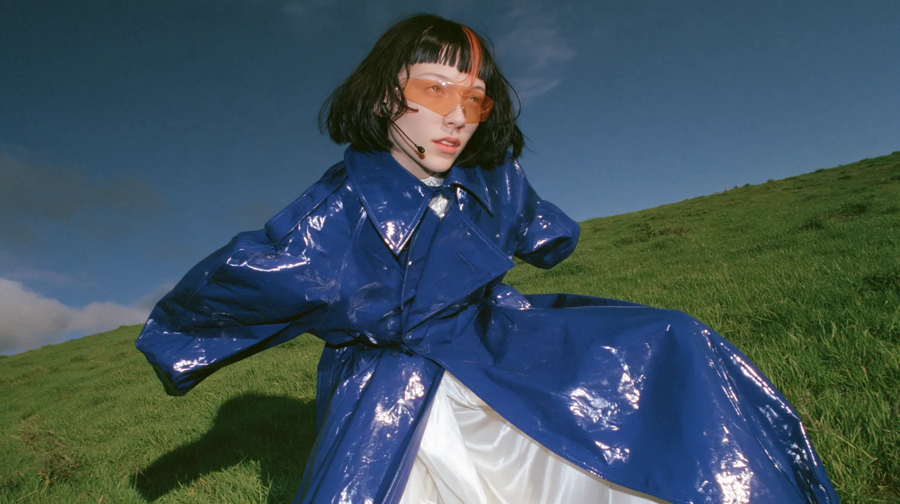 Y2K high fashion editorial, a 2001 couture magazine story: low wide shot of a young Caucasian American woman with pale skin and a glossy black blunt bob with heavy straight bangs and one clay-orange streak, a thin…