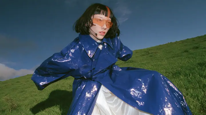Y2K high fashion editorial, a 2001 couture magazine story: low wide shot of a young Caucasian American woman with pale skin and a glossy black blunt bob with heavy straight bangs and one clay-orange streak, a thin…