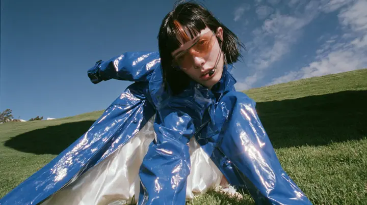 Y2K high fashion editorial, a 2001 couture magazine story: low wide shot of a young Caucasian American woman with pale skin and a glossy black blunt bob with heavy straight bangs and one clay-orange streak, a thin…