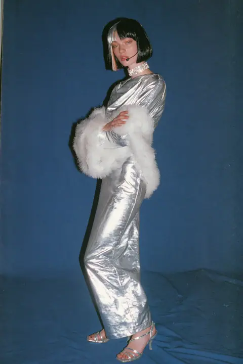 Y2K high fashion editorial, a 2001 couture magazine story: full-length shot, head to toe with the feet in frame, of a young Caucasian American woman with pale skin and a glossy black blunt bob with heavy straight…