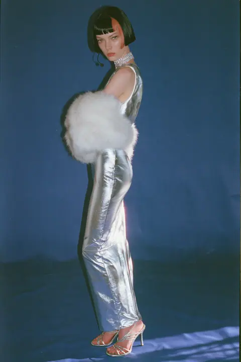 Y2K high fashion editorial, a 2001 couture magazine story: full-length shot, head to toe with the feet in frame, of a young Caucasian American woman with pale skin and a glossy black blunt bob with heavy straight…