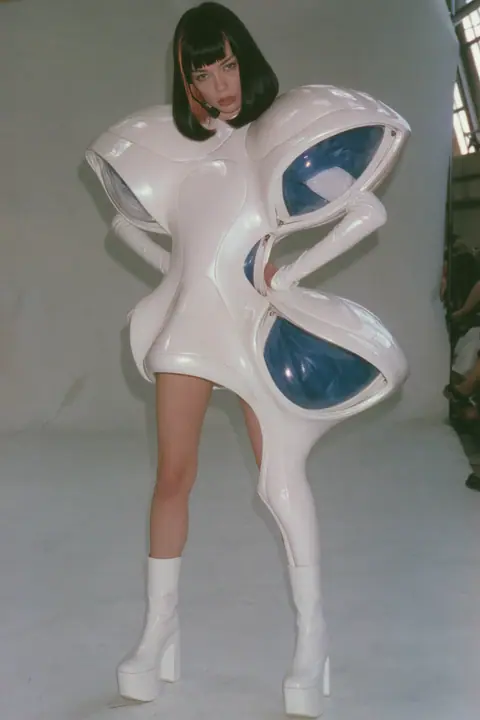 Y2K high fashion editorial, a 2001 couture magazine story: full-length shot, head to toe with the feet in frame, of a young Caucasian American woman with pale skin and a glossy black blunt bob with heavy straight…