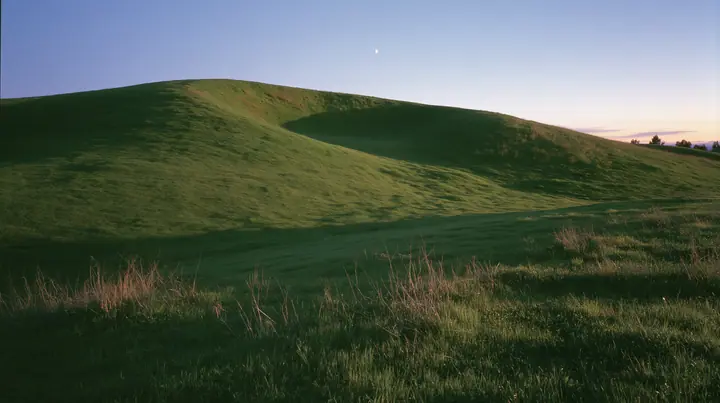 a smooth rolling bright-green grassy hill with soft curves, the grass short and velvety at dusk just after sunset, the grass dark green, a gradient sky from deep blue to soft lavender and amber at the horizon, the…