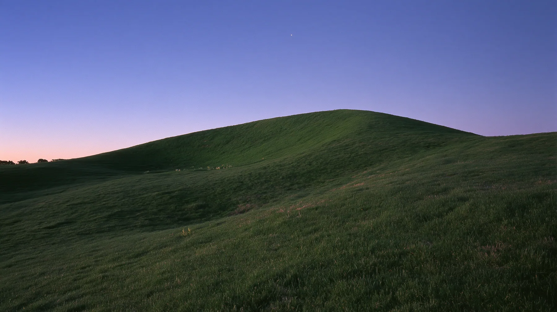 a smooth rolling bright-green grassy hill with soft curves, the grass short and velvety at dusk just after sunset, the grass dark green, a gradient sky from deep blue to soft lavender and amber at the horizon, the…