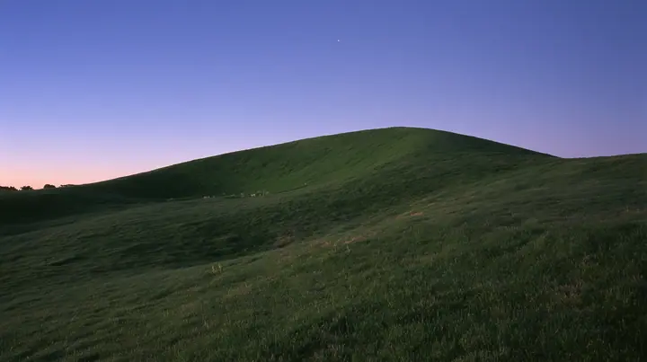 a smooth rolling bright-green grassy hill with soft curves, the grass short and velvety at dusk just after sunset, the grass dark green, a gradient sky from deep blue to soft lavender and amber at the horizon, the…