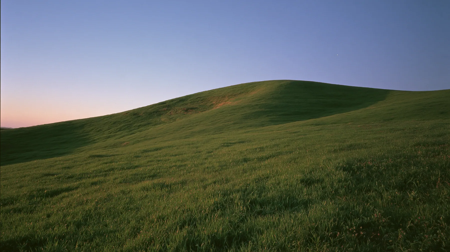 a smooth rolling bright-green grassy hill with soft curves, the grass short and velvety at dusk just after sunset, the grass dark green, a gradient sky from deep blue to soft lavender and amber at the horizon, the…