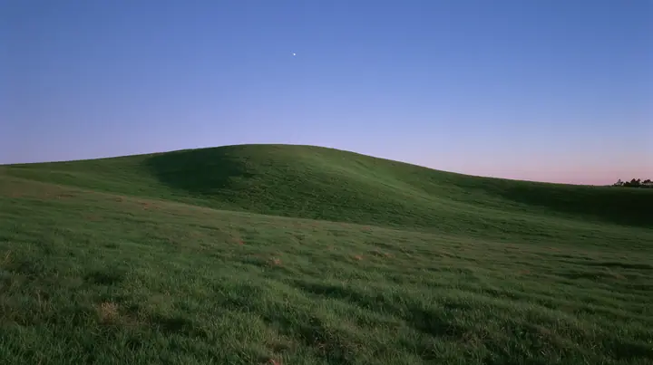 a smooth rolling bright-green grassy hill with soft curves, the grass short and velvety at dusk just after sunset, the grass dark green, a gradient sky from deep blue to soft lavender and amber at the horizon, the…