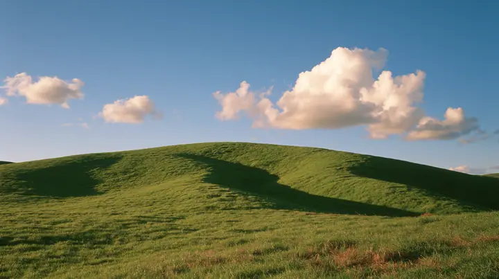 a smooth rolling bright-green grassy hill with soft curves, the grass short and velvety at golden hour, warm low sunlight raking across the grass, long soft shadows, a deep blue sky with glowing peach-edged cumulus,…