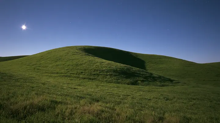 a smooth rolling bright-green grassy hill with soft curves, the grass short and velvety at night under a clear deep navy sky full of stars, a bright moon lighting the curve of the hill silver, calm, long exposure…