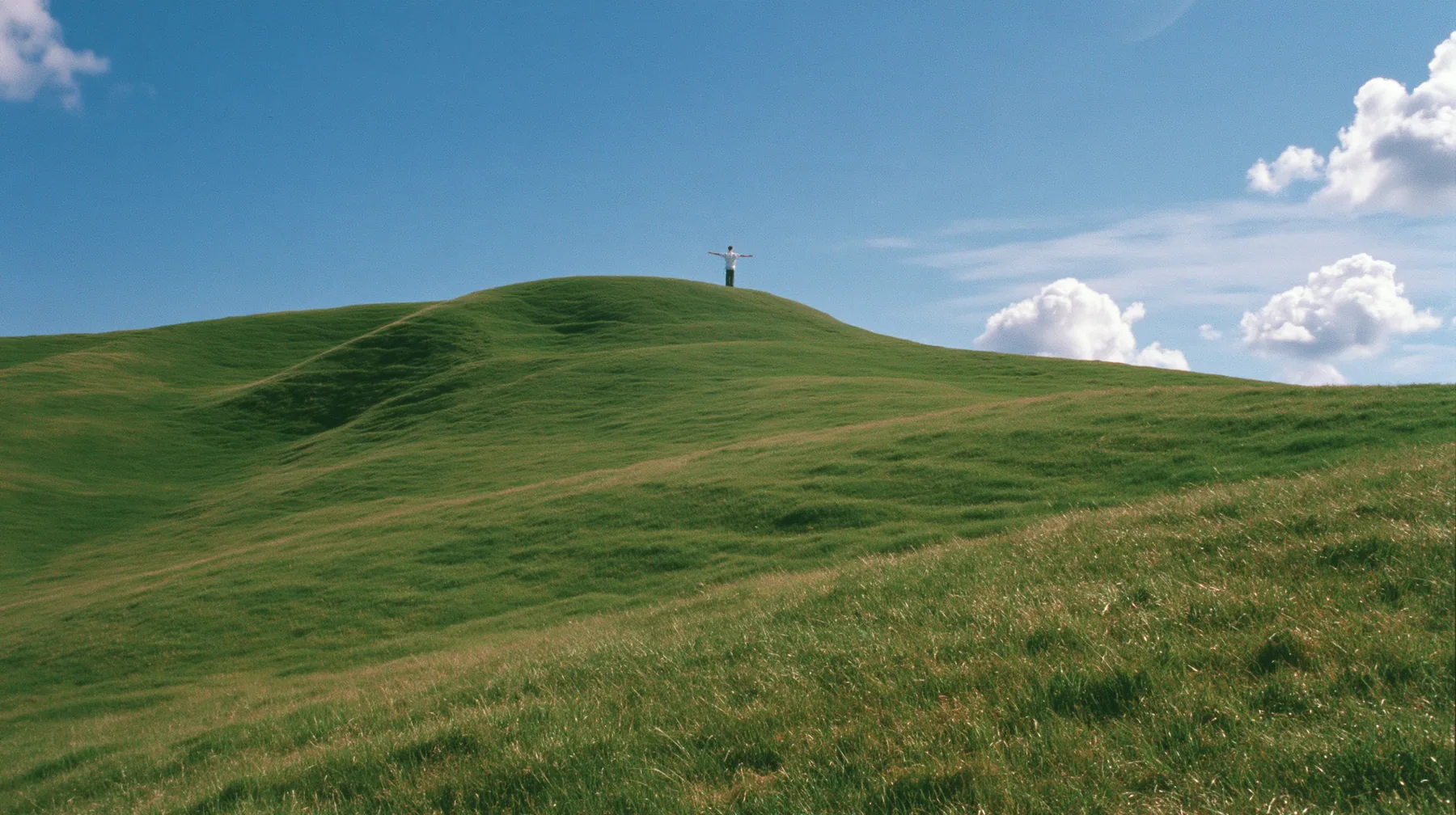 Y2K high fashion editorial photograph, 2001: a vast wide shot of a smooth rolling bright-green grassy hill with soft curves, the grass short and velvety with a tiny lone figure standing on the crest seen from behind,…