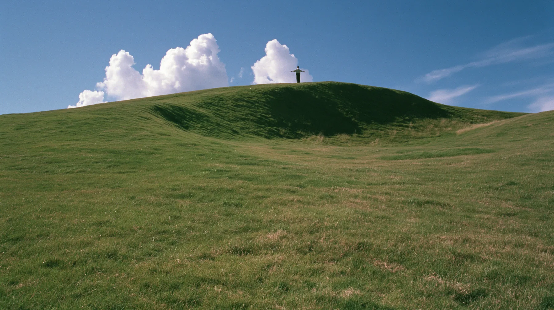 Y2K high fashion editorial photograph, 2001: a vast wide shot of a smooth rolling bright-green grassy hill with soft curves, the grass short and velvety with a tiny lone figure standing on the crest seen from behind,…
