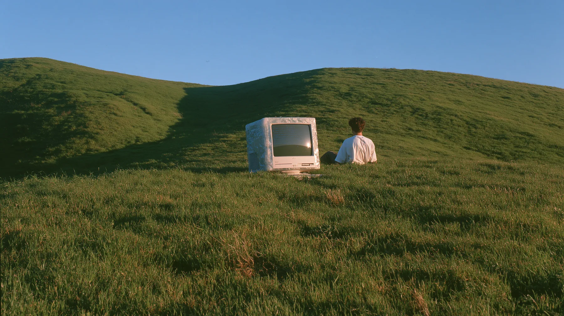 Y2K high fashion editorial photograph, 2001: a translucent ice-blue 2001 CRT computer monitor sitting alone on the crest of a smooth rolling bright-green grassy hill with soft curves, the grass short and velvety at…