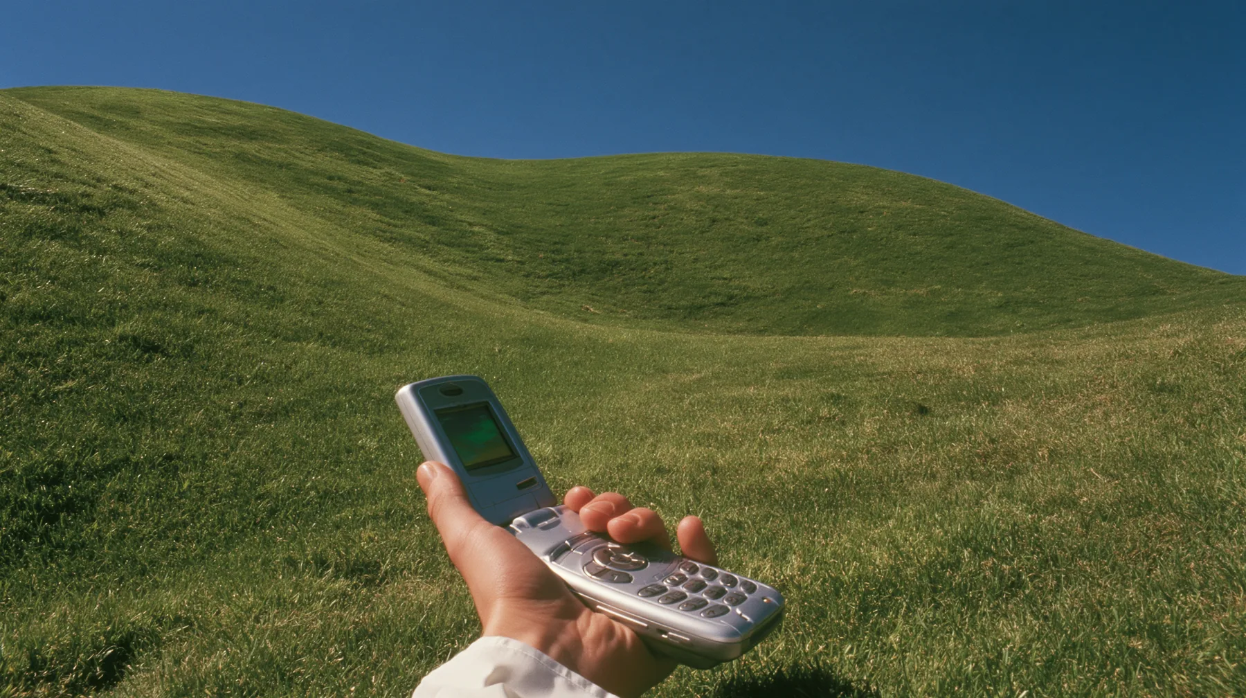 Y2K high fashion editorial photograph, 2001: first-person view of a hand holding an open silver flip phone against a smooth rolling bright-green grassy hill with soft curves, the grass short and velvety and a deep…