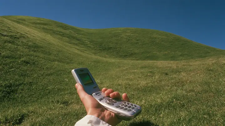 Y2K high fashion editorial photograph, 2001: first-person view of a hand holding an open silver flip phone against a smooth rolling bright-green grassy hill with soft curves, the grass short and velvety and a deep…