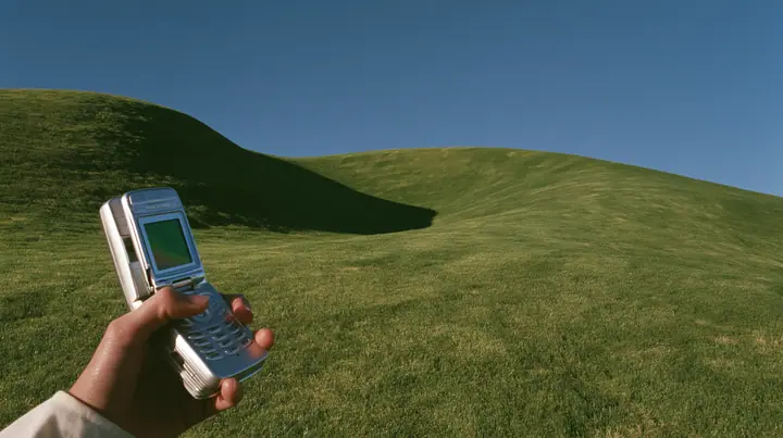 Y2K high fashion editorial photograph, 2001: first-person view of a hand holding an open silver flip phone against a smooth rolling bright-green grassy hill with soft curves, the grass short and velvety and a deep…