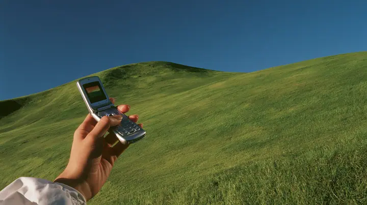 Y2K high fashion editorial photograph, 2001: first-person view of a hand holding an open silver flip phone against a smooth rolling bright-green grassy hill with soft curves, the grass short and velvety and a deep…