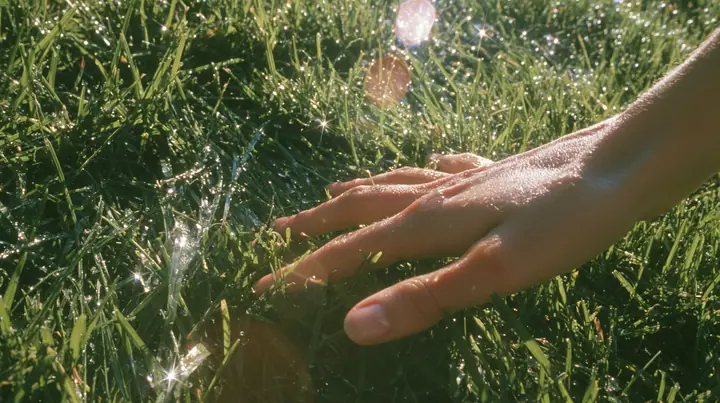 Y2K high fashion editorial photograph, 2001: first-person view of a hand gently brushing the tips of short bright-green grass, sunlight through the blades, dew, shallow depth of field, no text, no letters, no logos