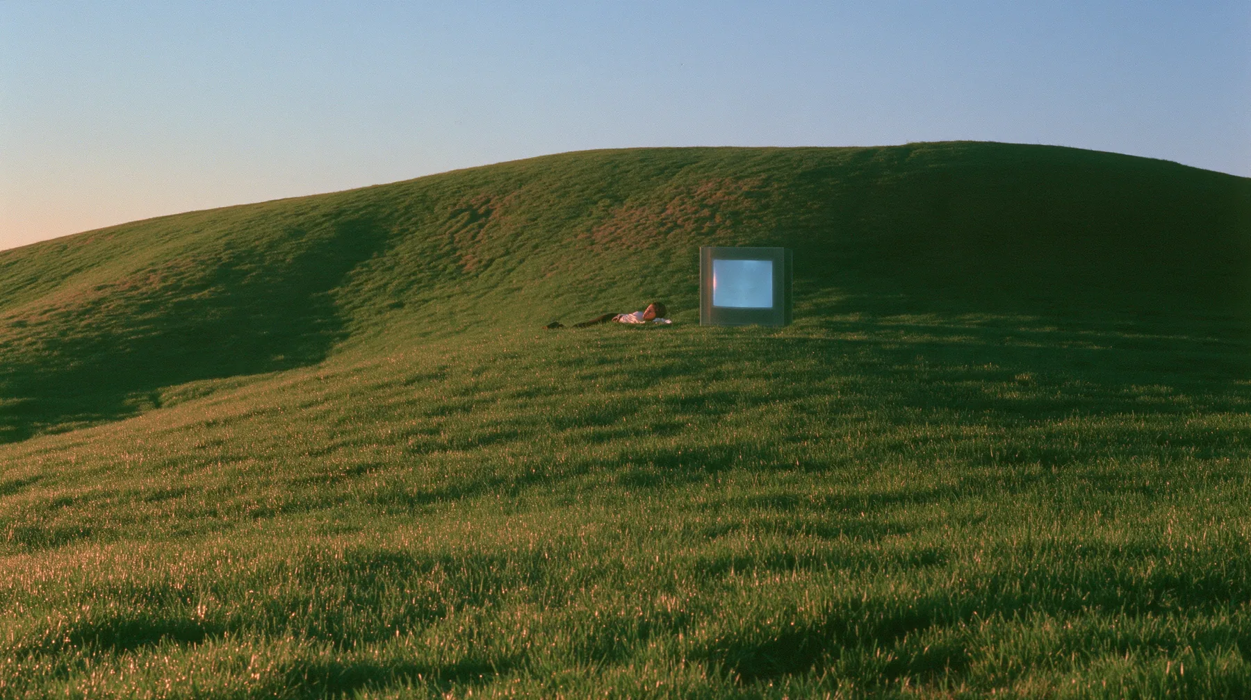 Y2K high fashion editorial photograph, 2001: a smooth rolling bright-green grassy hill with soft curves, the grass short and velvety at dusk, a dark translucent CRT monitor and a person lying in the grass beside it…