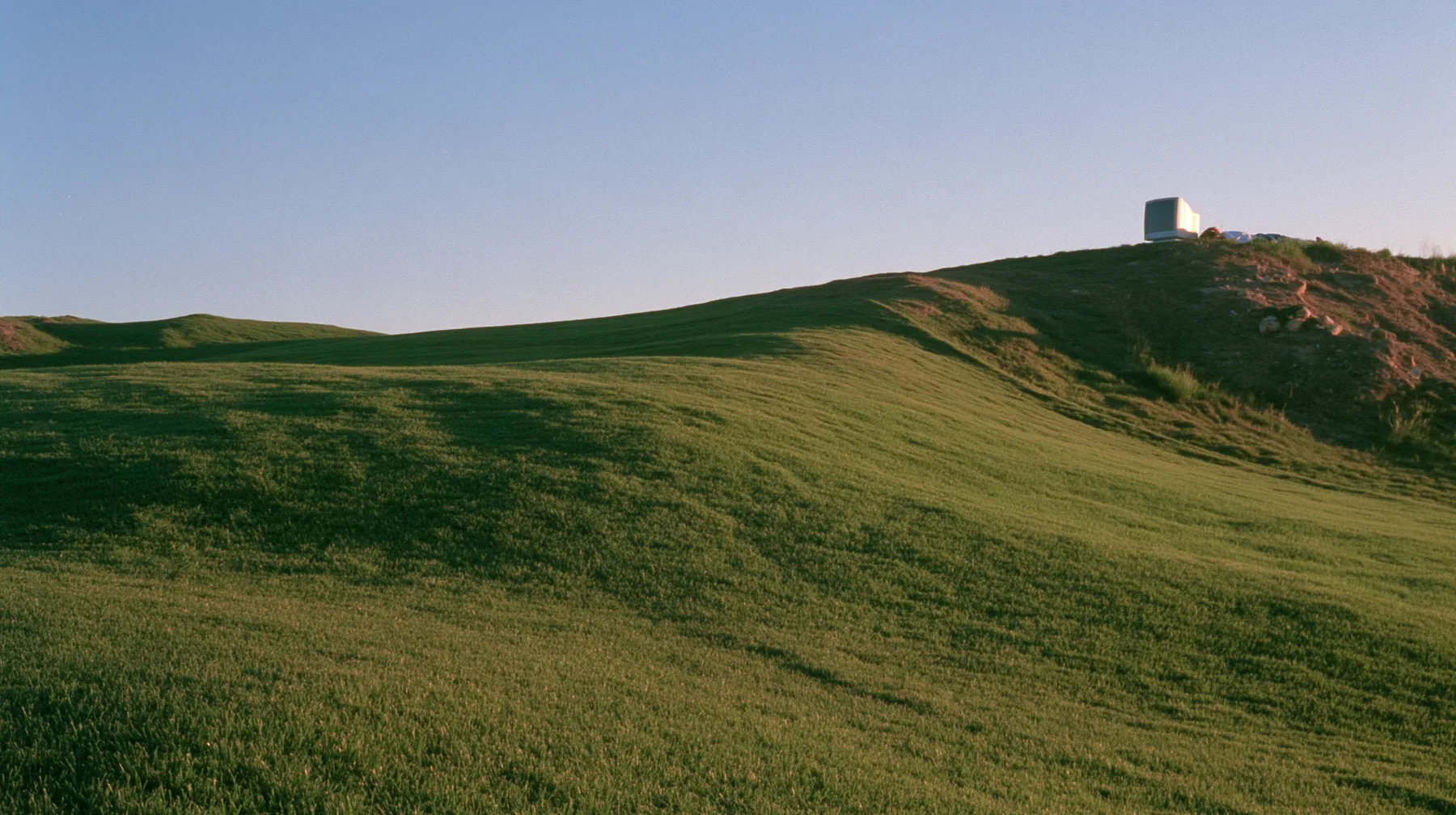 Y2K high fashion editorial photograph, 2001: a smooth rolling bright-green grassy hill with soft curves, the grass short and velvety at dusk, a dark translucent CRT monitor and a person lying in the grass beside it…