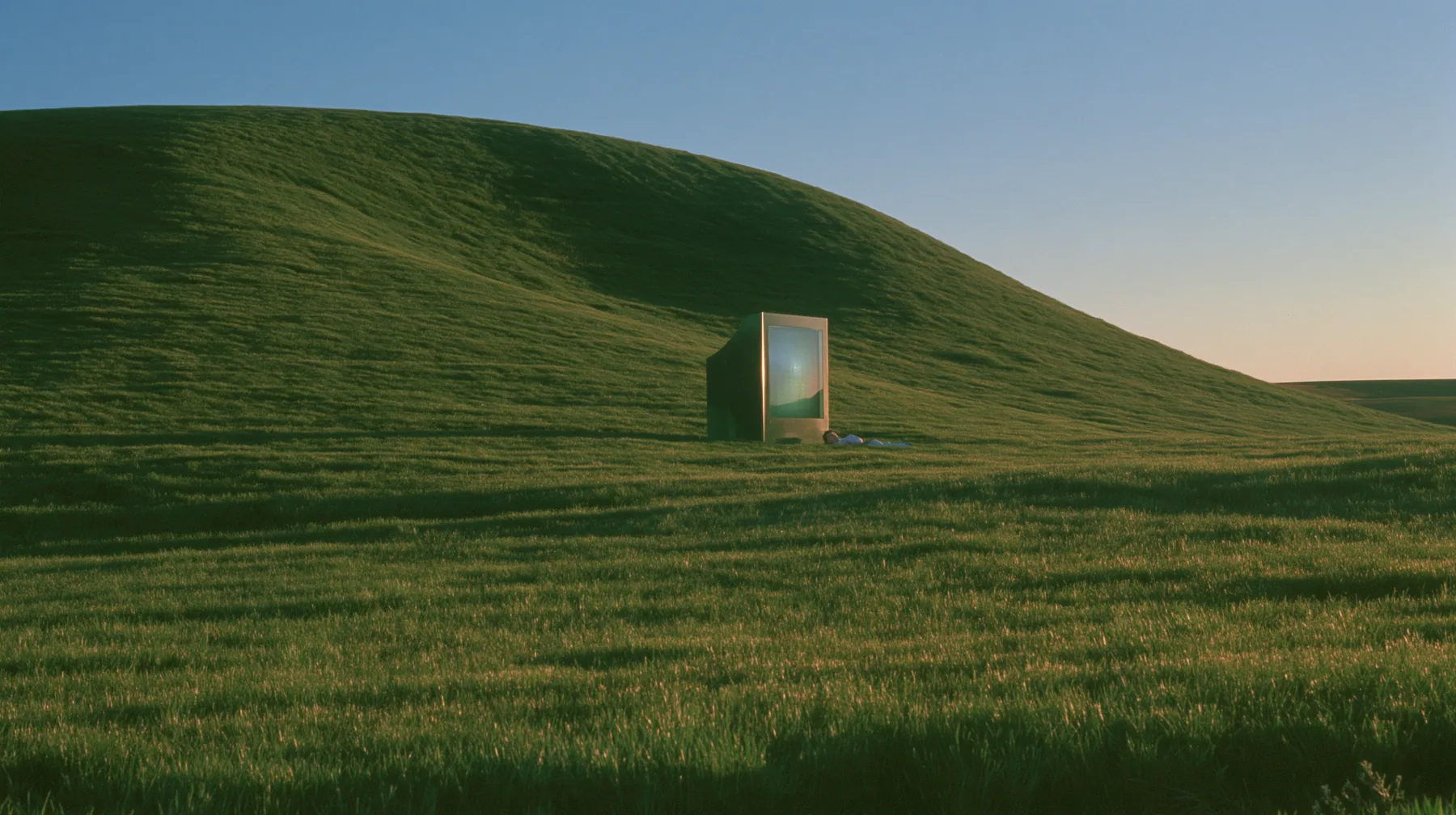 Y2K high fashion editorial photograph, 2001: a smooth rolling bright-green grassy hill with soft curves, the grass short and velvety at dusk, a dark translucent CRT monitor and a person lying in the grass beside it…