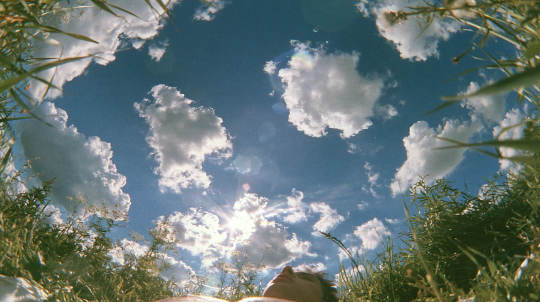 Y2K high fashion editorial photograph, 2001: first-person view lying on your back in the grass looking straight up at a deep blue sky with soft white cumulus clouds, grass blades at the edges of frame, bright sun, no…