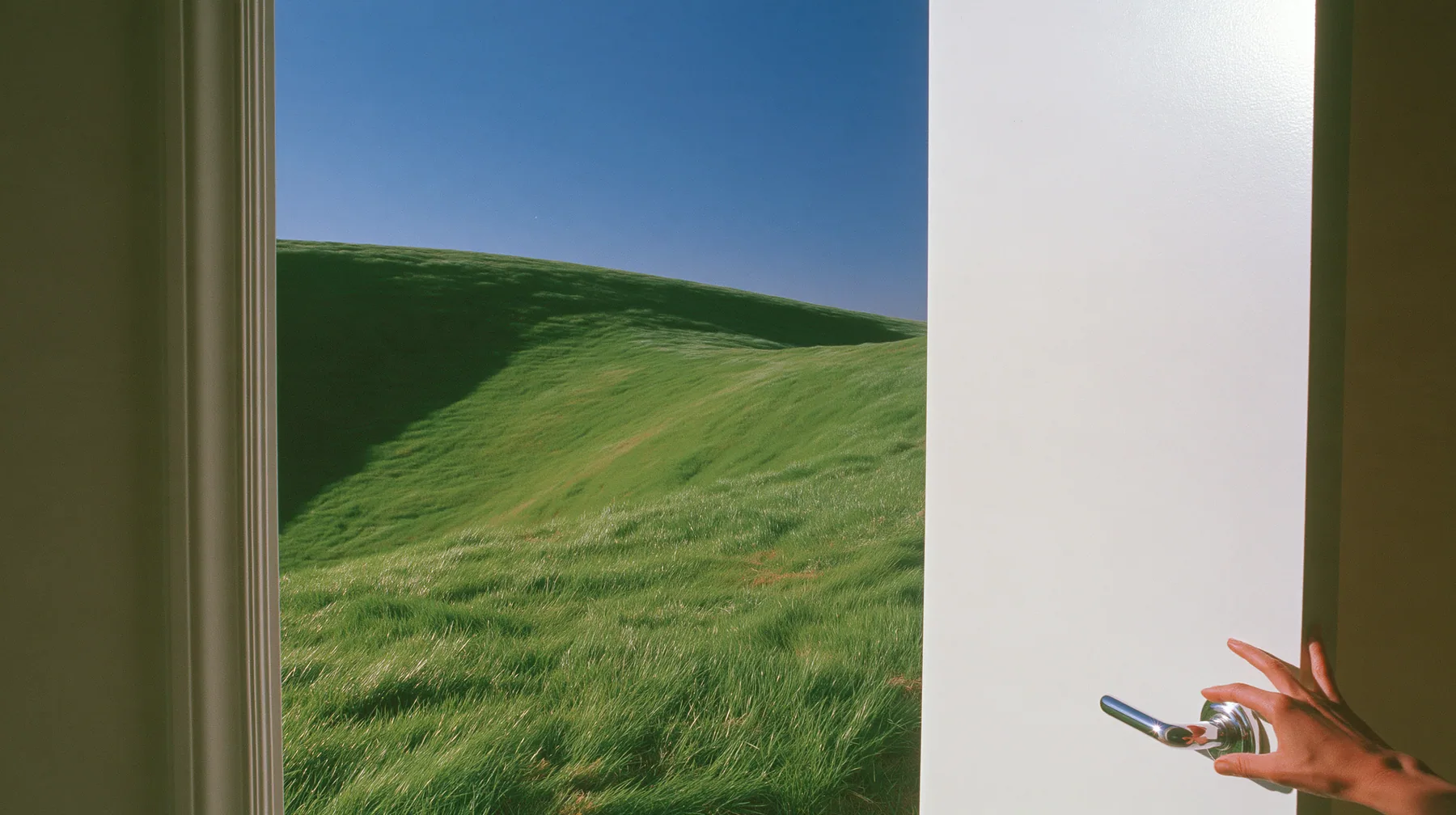 Y2K high fashion editorial photograph, 2001: first-person view of a hand on the chrome handle of a tall white door swinging open onto a bright morning, a smooth rolling bright-green grassy hill with soft curves, the…