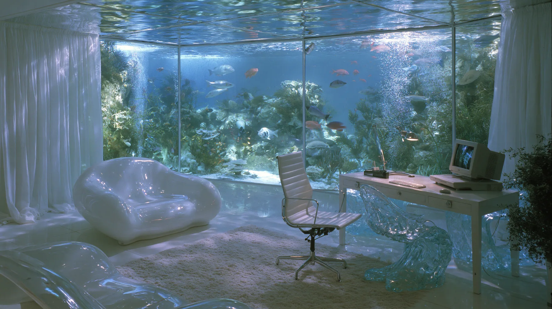 Y2K high fashion editorial photograph, 2001: a vivid tropical aquarium wall glowing in a 2001 high-fashion minimalist white loft apartment with molded white plastic furniture, a white lacquer desk, chrome and…