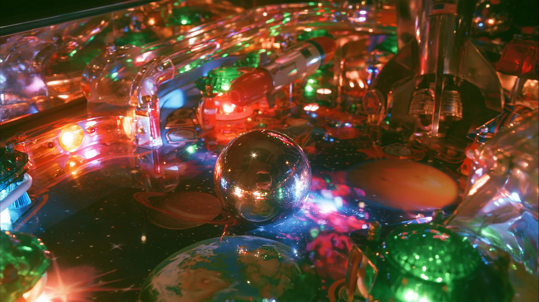 Y2K high fashion editorial photograph, 2001: a luxury product photograph of a glossy chrome and candy-colored space-themed pinball table, a shiny silver ball, glowing bumpers, a rocket ramp and planets, no text, no…