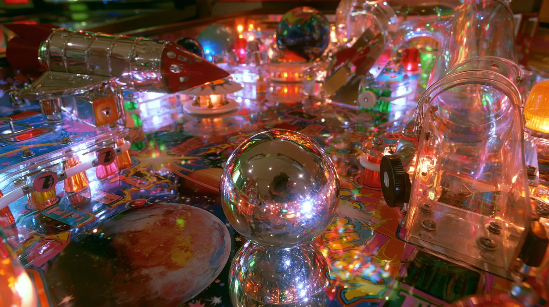 Y2K high fashion editorial photograph, 2001: a luxury product photograph of a glossy chrome and candy-colored space-themed pinball table, a shiny silver ball, glowing bumpers, a rocket ramp and planets, no text, no…