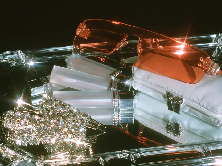 Y2K high fashion editorial photograph, 2001: a luxury beauty still life of frosted lip-gloss tubes, rimless orange-tinted shield sunglasses, rhinestone hair clips and a tiny white baguette bag on a mirrored tray,…