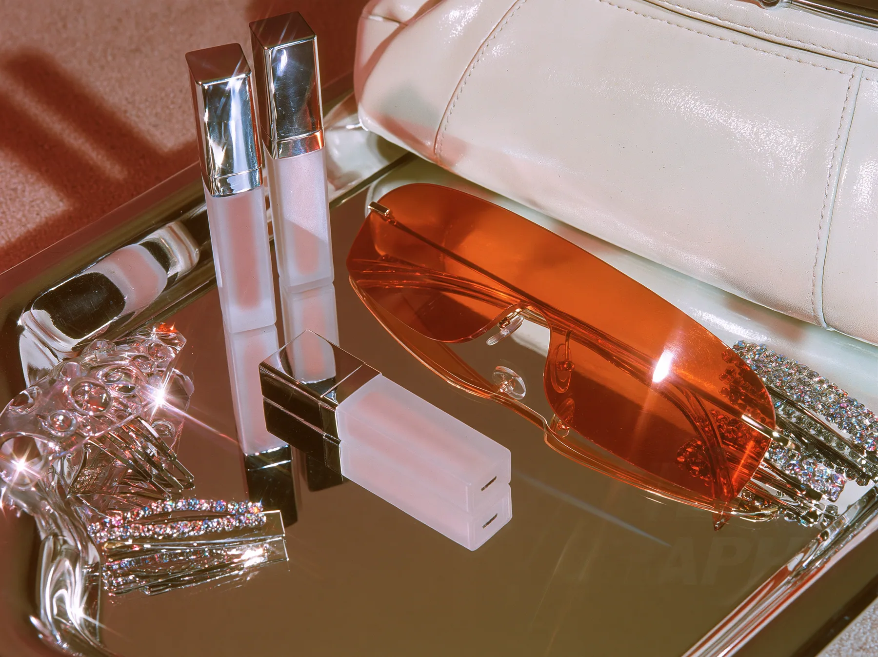 Y2K high fashion editorial photograph, 2001: a luxury beauty still life of frosted lip-gloss tubes, rimless orange-tinted shield sunglasses, rhinestone hair clips and a tiny white baguette bag on a mirrored tray,…