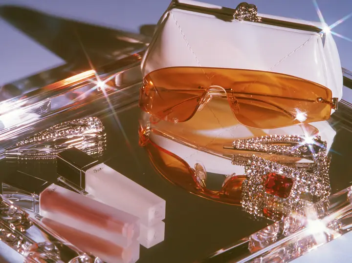 Y2K high fashion editorial photograph, 2001: a luxury beauty still life of frosted lip-gloss tubes, rimless orange-tinted shield sunglasses, rhinestone hair clips and a tiny white baguette bag on a mirrored tray,…