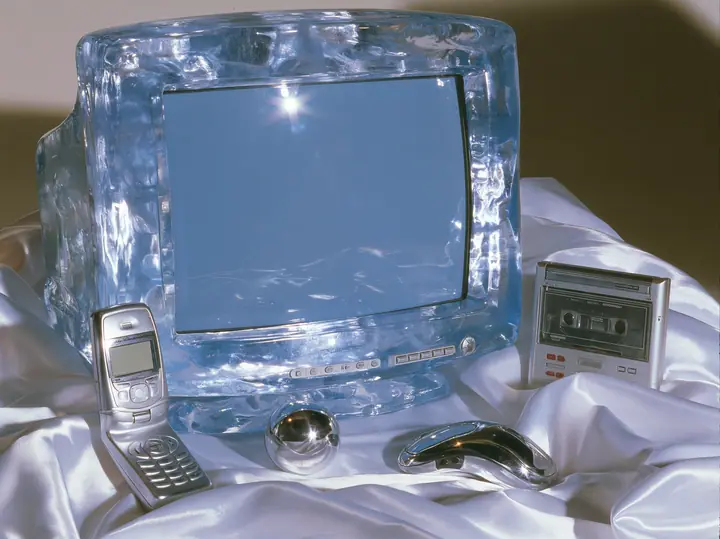 Y2K high fashion editorial photograph, 2001: a luxury still life of a translucent ice-blue CRT monitor, a silver flip phone, a chrome ball mouse and a portable CD player arranged on white satin, glossy studio light,…