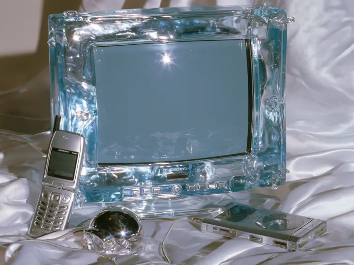 Y2K high fashion editorial photograph, 2001: a luxury still life of a translucent ice-blue CRT monitor, a silver flip phone, a chrome ball mouse and a portable CD player arranged on white satin, glossy studio light,…