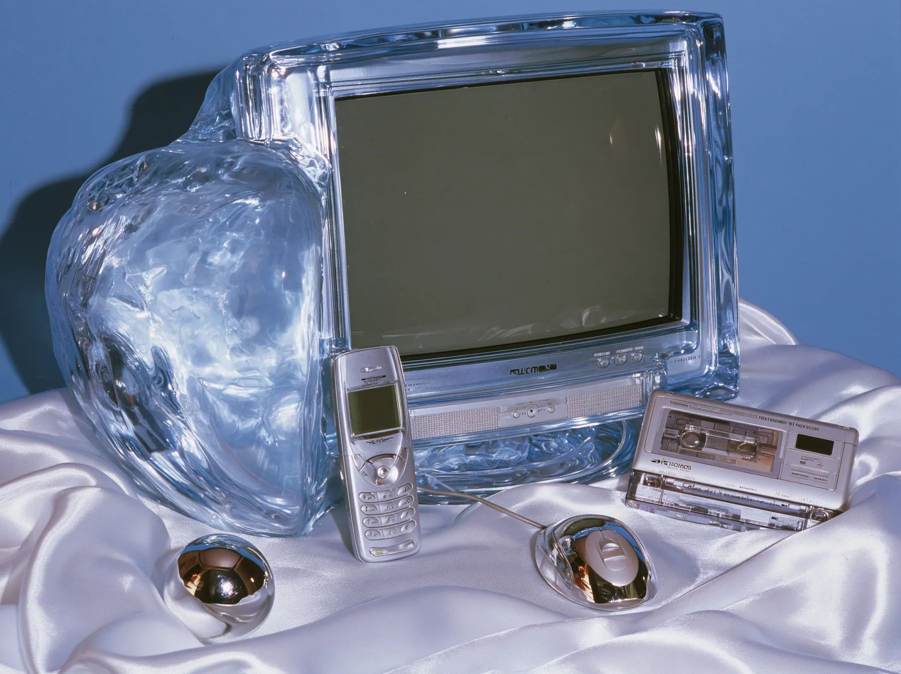 Y2K high fashion editorial photograph, 2001: a luxury still life of a translucent ice-blue CRT monitor, a silver flip phone, a chrome ball mouse and a portable CD player arranged on white satin, glossy studio light,…