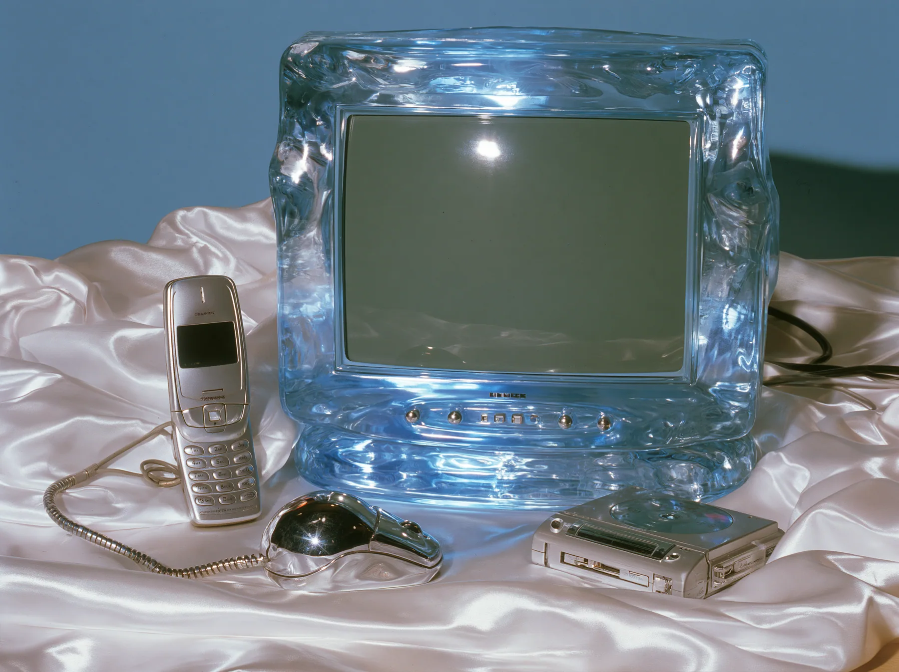 Y2K high fashion editorial photograph, 2001: a luxury still life of a translucent ice-blue CRT monitor, a silver flip phone, a chrome ball mouse and a portable CD player arranged on white satin, glossy studio light,…