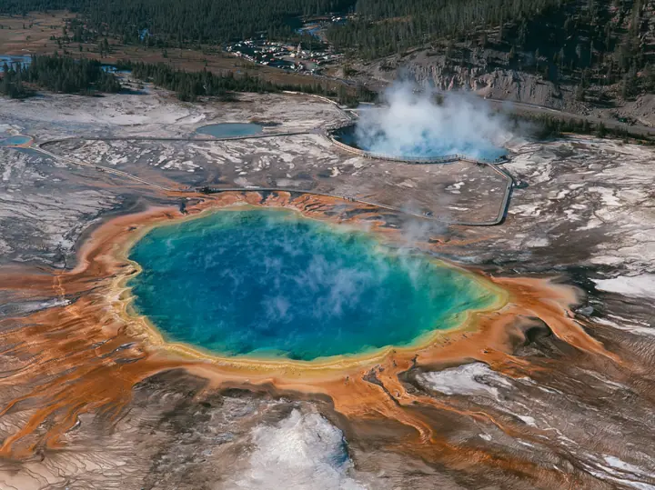 Y2K high fashion editorial photograph, 2001: a luxury travel campaign aerial of a vast hot spring in rings of vivid blue, green, yellow and orange, steam drifting, a boardwalk at its edge, Yellowstone-like, no text,…