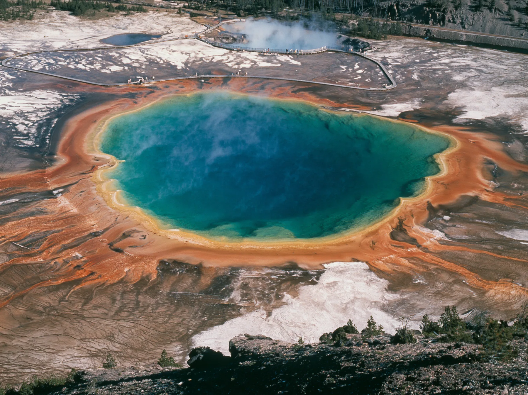 Y2K high fashion editorial photograph, 2001: a luxury travel campaign aerial of a vast hot spring in rings of vivid blue, green, yellow and orange, steam drifting, a boardwalk at its edge, Yellowstone-like, no text,…