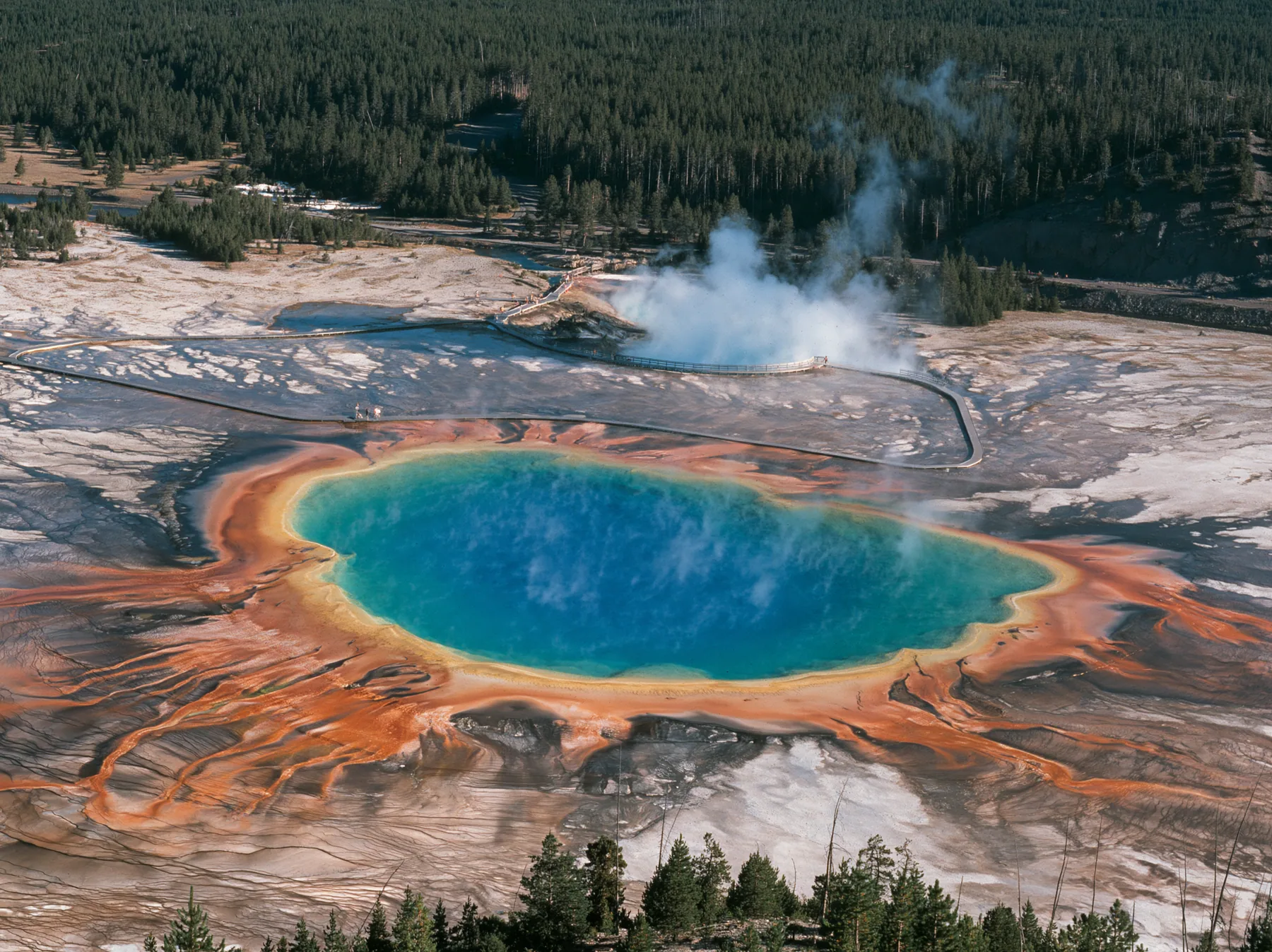 Y2K high fashion editorial photograph, 2001: a luxury travel campaign aerial of a vast hot spring in rings of vivid blue, green, yellow and orange, steam drifting, a boardwalk at its edge, Yellowstone-like, no text,…