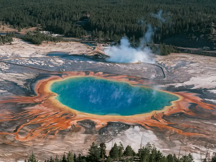 Y2K high fashion editorial photograph, 2001: a luxury travel campaign aerial of a vast hot spring in rings of vivid blue, green, yellow and orange, steam drifting, a boardwalk at its edge, Yellowstone-like, no text,…