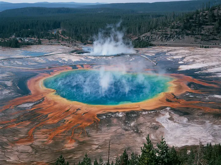Y2K high fashion editorial photograph, 2001: a luxury travel campaign aerial of a vast hot spring in rings of vivid blue, green, yellow and orange, steam drifting, a boardwalk at its edge, Yellowstone-like, no text,…