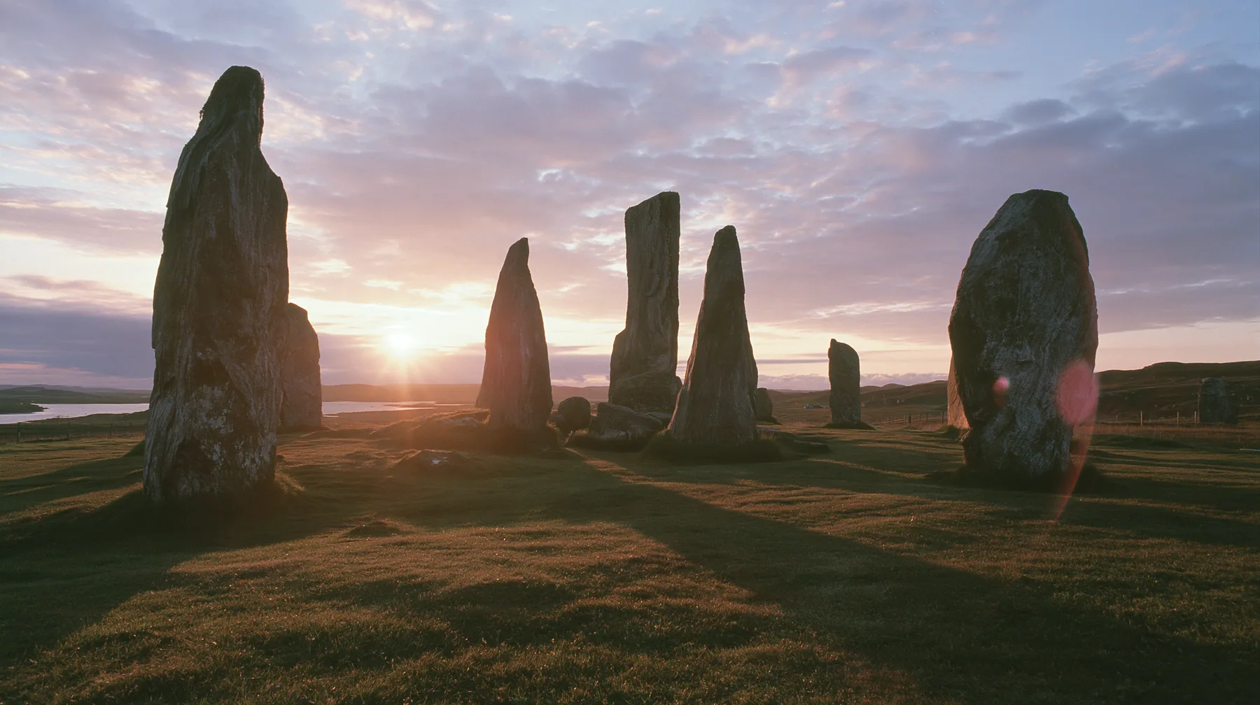 Y2K high fashion editorial photograph, 2001: a luxury campaign landscape of ancient standing stones silhouetted against a glowing violet and amber sunset, medium-format slide film, no text, no letters, no logos