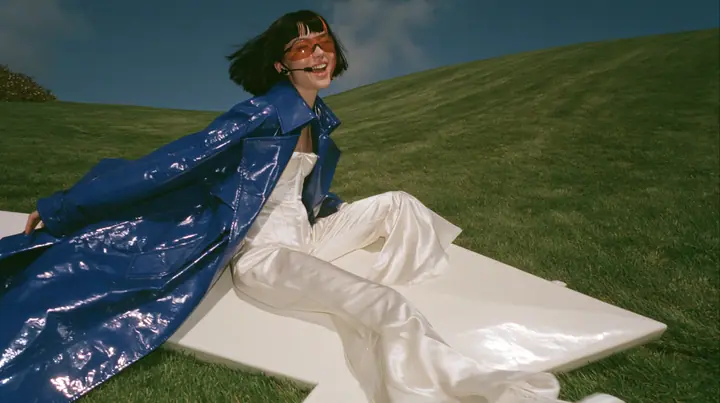 Y2K high fashion editorial, a 2001 couture magazine story: a wide shot of a young Caucasian American woman with pale skin and a glossy black blunt bob with heavy straight bangs and one clay-orange streak, a thin…