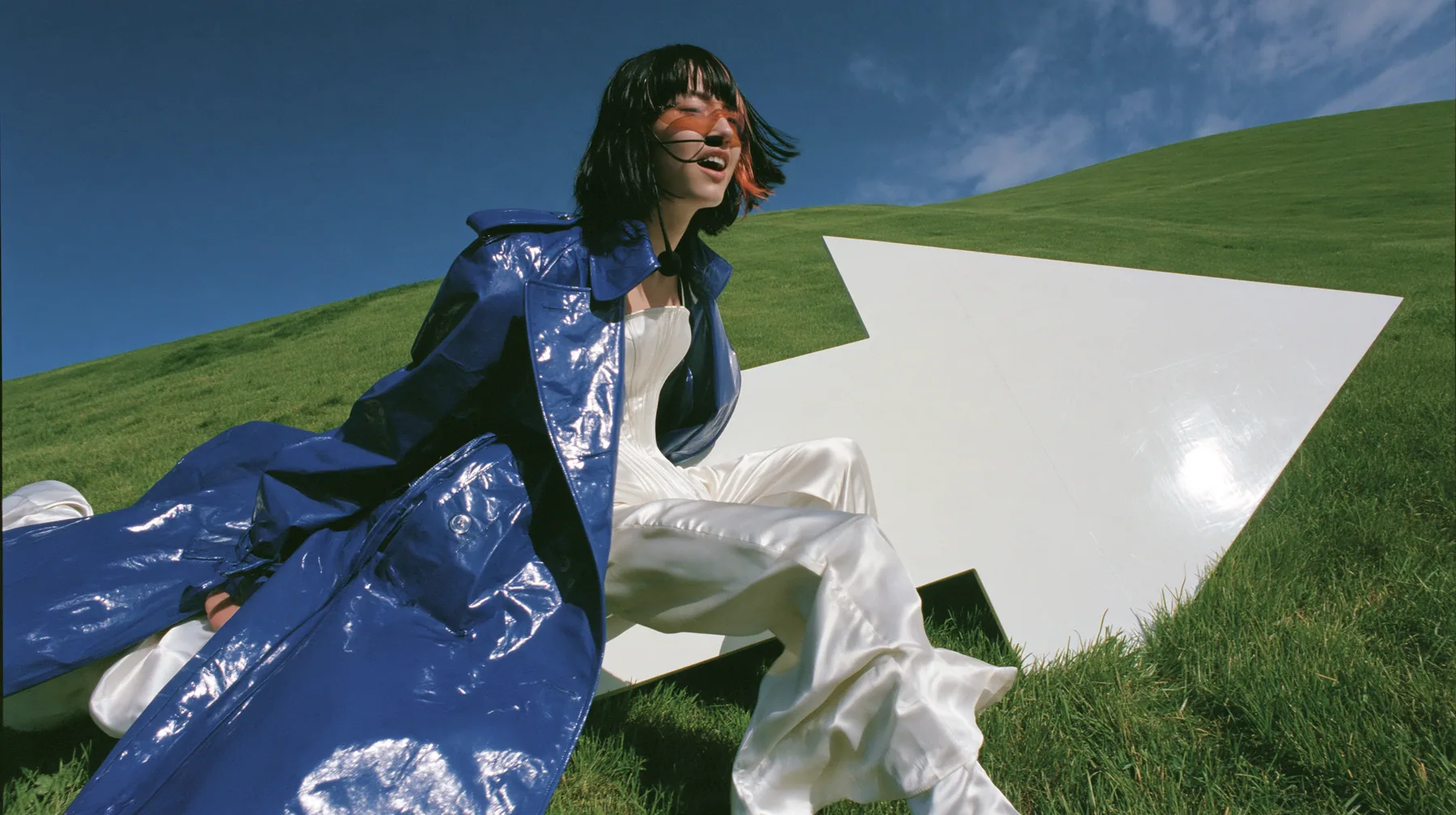 Y2K high fashion editorial, a 2001 couture magazine story: a wide shot of a young Caucasian American woman with pale skin and a glossy black blunt bob with heavy straight bangs and one clay-orange streak, a thin…