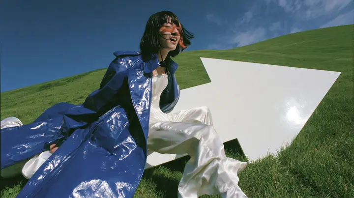 Y2K high fashion editorial, a 2001 couture magazine story: a wide shot of a young Caucasian American woman with pale skin and a glossy black blunt bob with heavy straight bangs and one clay-orange streak, a thin…