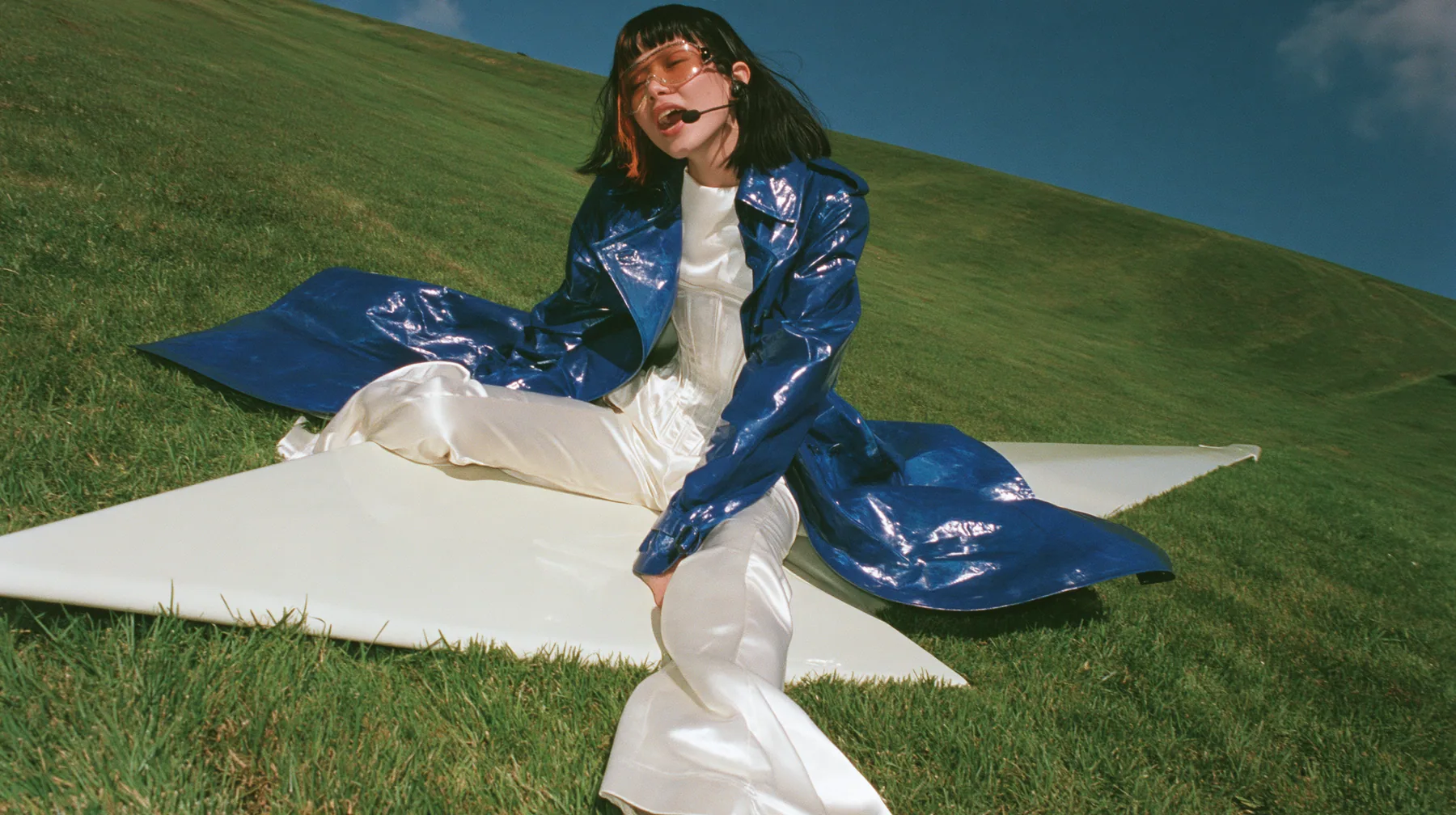 Y2K high fashion editorial, a 2001 couture magazine story: a wide shot of a young Caucasian American woman with pale skin and a glossy black blunt bob with heavy straight bangs and one clay-orange streak, a thin…