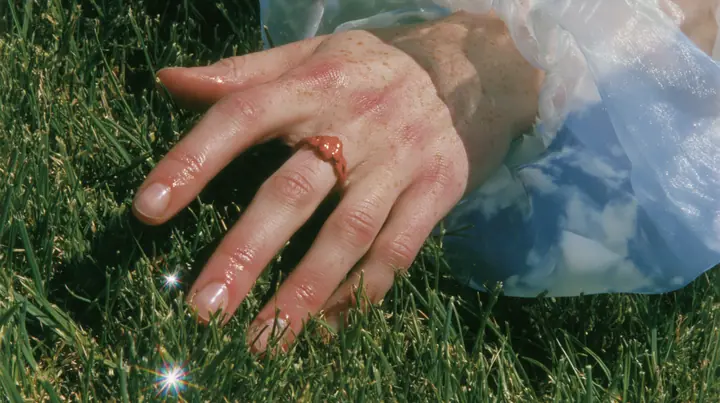 Y2K high fashion editorial, a 2001 couture magazine story: a macro of a pale freckled hand with a small clay-orange enamel ring brushing the tips of short bright-green grass, a sleeve of sky-print chiffon, sunlight…