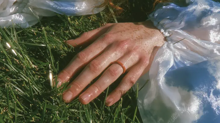 Y2K high fashion editorial, a 2001 couture magazine story: a macro of a pale freckled hand with a small clay-orange enamel ring brushing the tips of short bright-green grass, a sleeve of sky-print chiffon, sunlight…